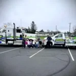 A group of children and drivers standing in a parking lot in front of two Bill's Towing trucks during a community event in Roseburg.