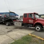 A red Bill's Towing flatbed truck loading a disabled dark minivan onto the bed in front of a blue commercial building in Winston, Oregon.
