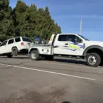 A white Bill's Towing truck towing a white Jeep SUV across a parking lot in Roseburg, Oregon.